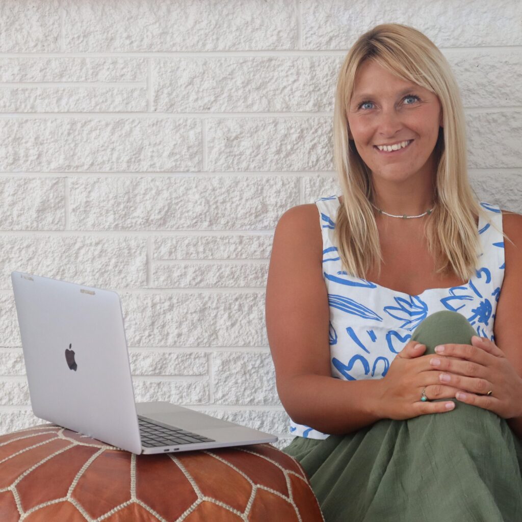 Katy Guildford - a business strategy & performance expert in New Zealand sat against a white brick wall with a laptop beside her.